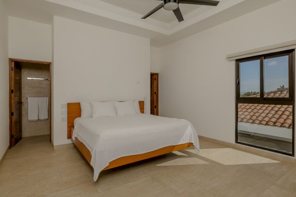 A modern bedroom with a large bed, ceiling fan, attached bathroom, and a window with a view of a tiled roof and greenery outside.