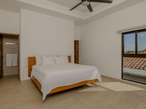 A modern bedroom with a large bed, ceiling fan, attached bathroom, and a window with a view of a tiled roof and greenery outside.