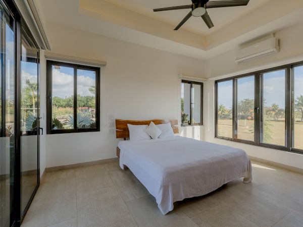 A bright bedroom with large windows, a double bed, white linens, a ceiling fan, and tiled floors. The room has a peaceful, airy ambiance.
