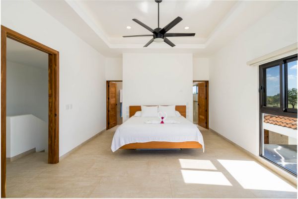 A spacious, bright bedroom with a modern ceiling fan, large bed, wooden doors, and a window showing an outdoor view in natural light.