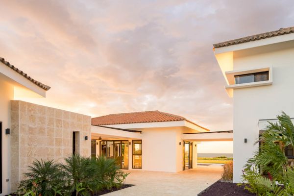 A modern house with a clean, open courtyard, light-colored plaster and stone walls, and landscaped greenery under a dramatic, cloudy sky.