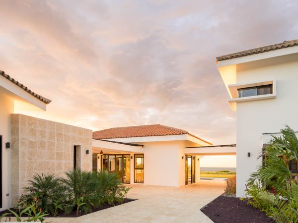 A modern house with a clean, open courtyard, light-colored plaster and stone walls, and landscaped greenery under a dramatic, cloudy sky.