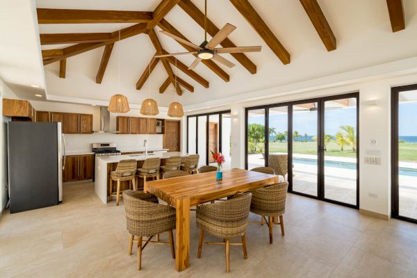 A modern kitchen and dining area with wooden beams, a large table, wicker chairs, and pendant lights. Glass doors open to a scenic outdoor view.