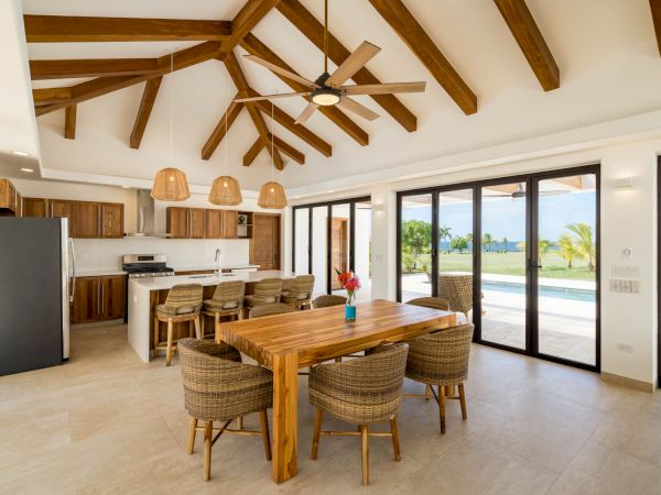 A modern kitchen and dining area with wooden beams, a large table, wicker chairs, and pendant lights. Glass doors open to a scenic outdoor view.