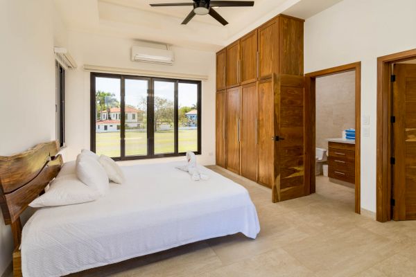 A bedroom featuring a large bed with white linens, wooden wardrobes, a ceiling fan, and an en-suite bathroom. Large windows offer a view outside.