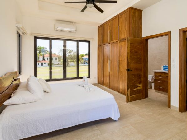 A bedroom featuring a large bed with white linens, wooden wardrobes, a ceiling fan, and an en-suite bathroom. Large windows offer a view outside.