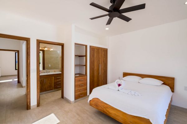 A modern bedroom featuring a wooden bed, a ceiling fan, and an open bathroom area with wooden cabinets and a vanity. The space appears clean and bright.