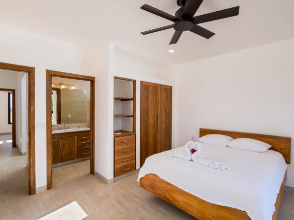 A modern bedroom featuring a wooden bed, a ceiling fan, and an open bathroom area with wooden cabinets and a vanity. The space appears clean and bright.