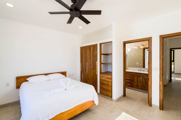 A bright bedroom with a modern wooden bed, white linens, a ceiling fan, a wardrobe, and an ensuite bathroom with a wooden vanity, ending the sentence.