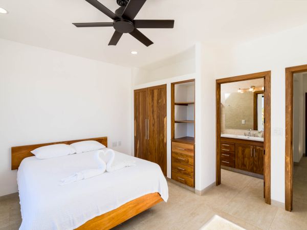 A bright bedroom with a modern wooden bed, white linens, a ceiling fan, a wardrobe, and an ensuite bathroom with a wooden vanity, ending the sentence.