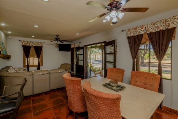 A living room with a dining table and four orange chairs, couch, ceiling fans, TV, open French doors, and windows with curtains.