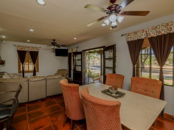 A living room with a dining table and four orange chairs, couch, ceiling fans, TV, open French doors, and windows with curtains.