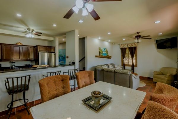 The image shows an open-plan living area with a dining table, kitchen, and a cozy corner sofa, adorned with warm lighting and neutral decor.