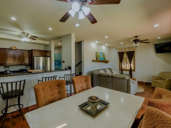 The image shows an open-plan living area with a dining table, kitchen, and a cozy corner sofa, adorned with warm lighting and neutral decor.