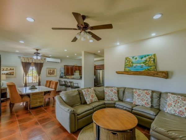 A cozy living room with a sectional sofa, a round coffee table, dining area, kitchen, ceiling fans, and wall art, all under warm recessed lighting.