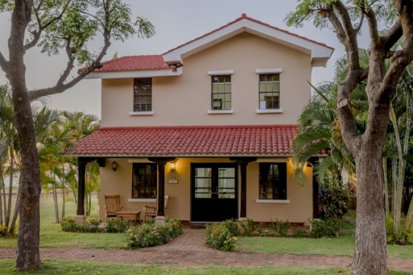 A two-story house with a red-tiled roof and beige exterior, surrounded by trees and greenery. The front porch has a wooden bench and warm lighting.