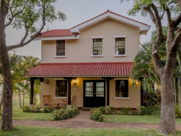 A two-story house with a red-tiled roof and beige exterior, surrounded by trees and greenery. The front porch has a wooden bench and warm lighting.