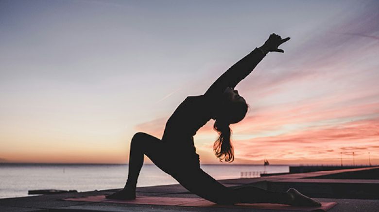 A person practices yoga in a lunge position on a rooftop at sunset, with the sky displaying vibrant hues.