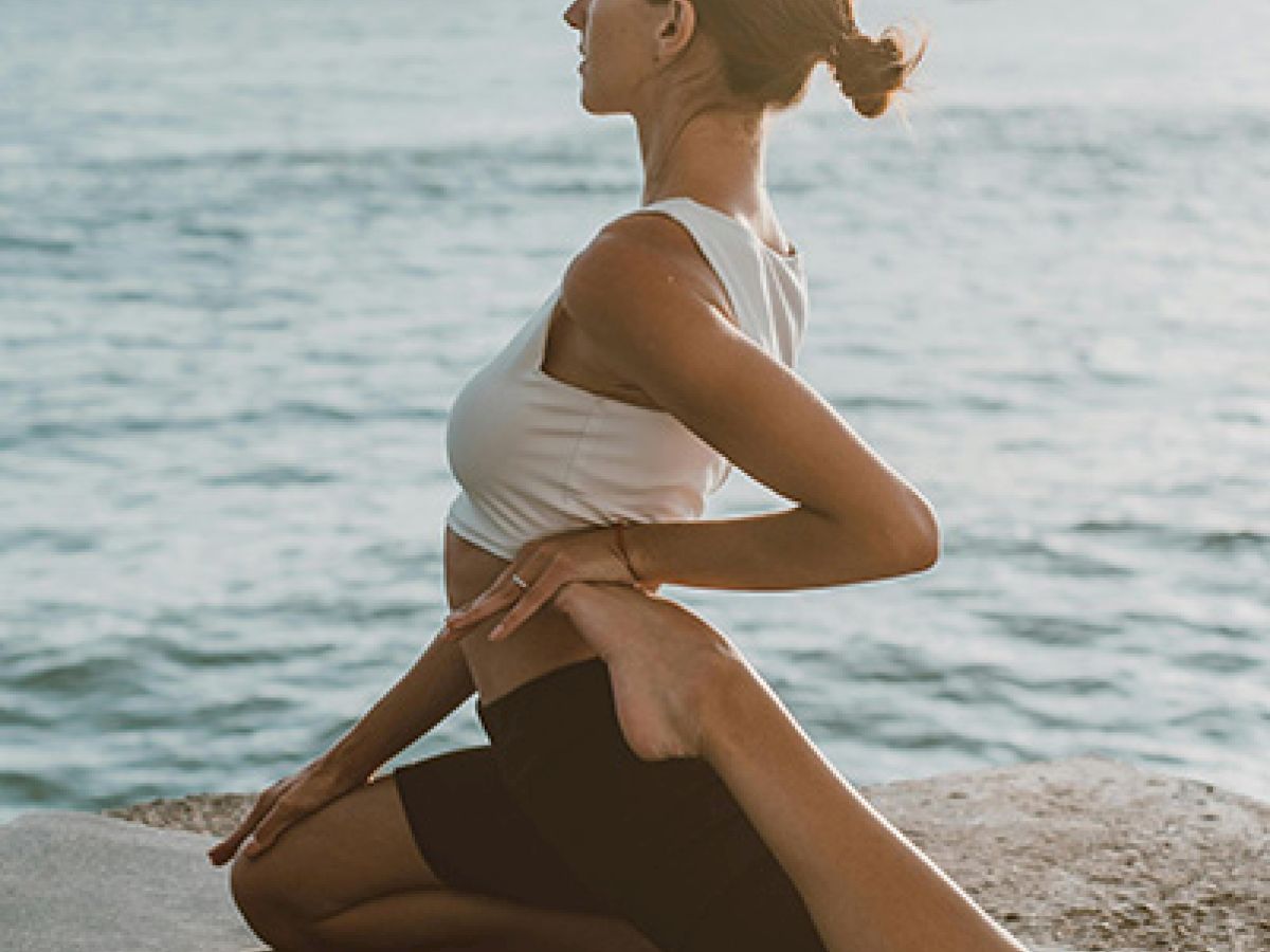 A person is performing a yoga pose on a mat near a body of water, with their hands clasped behind their back and head turned to the side.