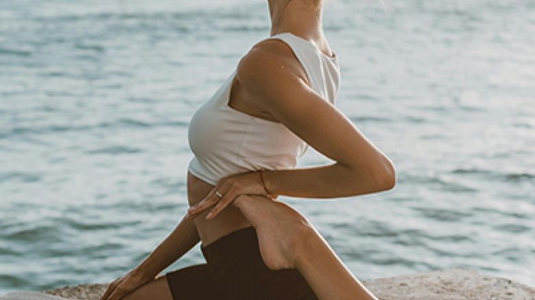 A person is performing a yoga pose on a mat near a body of water, with their hands clasped behind their back and head turned to the side.