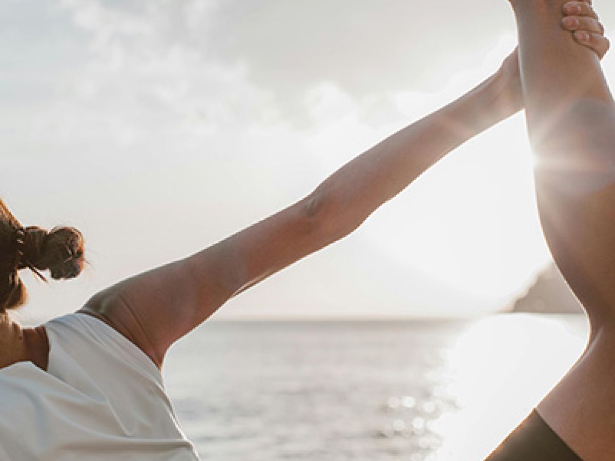 A person practicing yoga on a beach at sunset or sunrise, holding one leg up with their hand, dressed in a white top and black shorts, calm water behind.