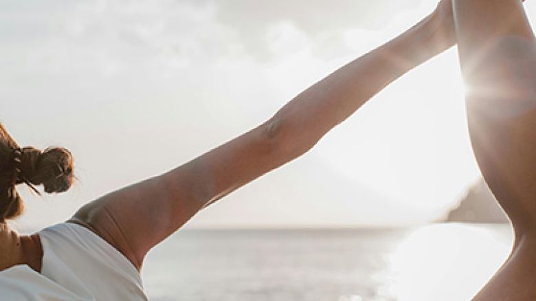 A person practicing yoga on a beach at sunset or sunrise, holding one leg up with their hand, dressed in a white top and black shorts, calm water behind.