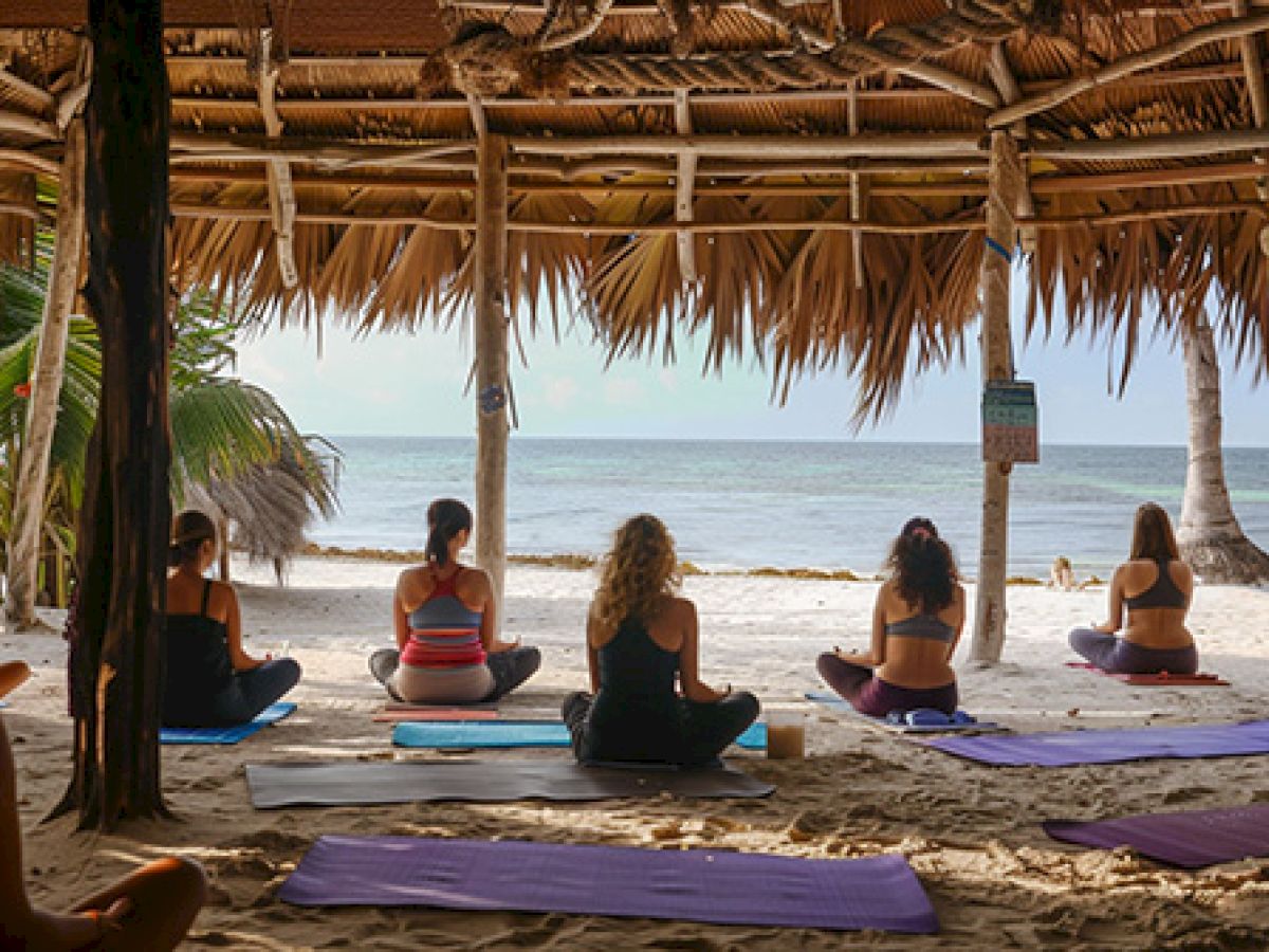 People are practicing yoga on a beach under a thatched roof structure, facing the ocean, surrounded by palm trees.