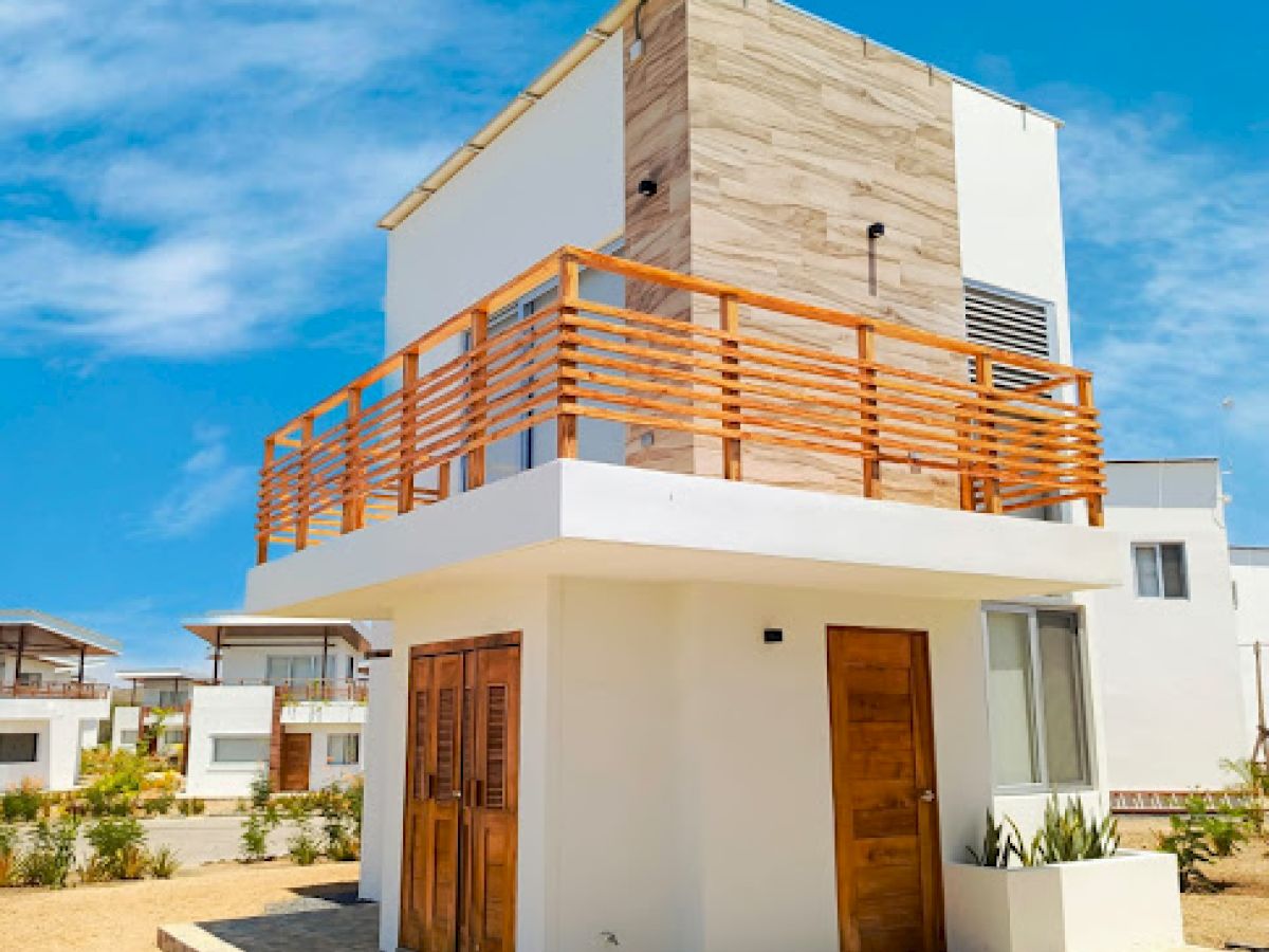 The image shows a modern two-story house with white walls, wooden accents, and a balcony on the upper floor, under a clear blue sky.