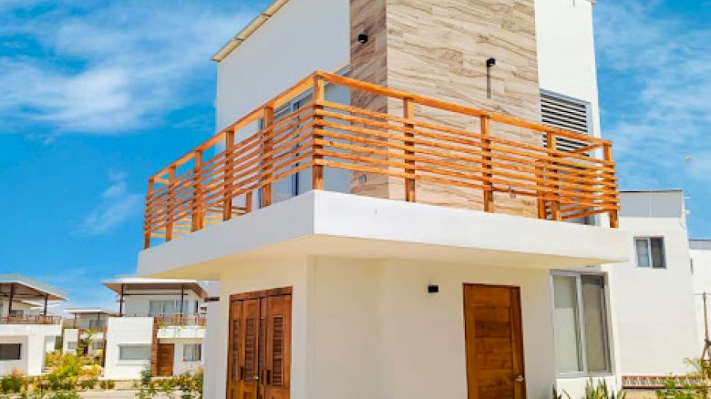 The image shows a modern two-story house with white walls, wooden accents, and a balcony on the upper floor, under a clear blue sky.