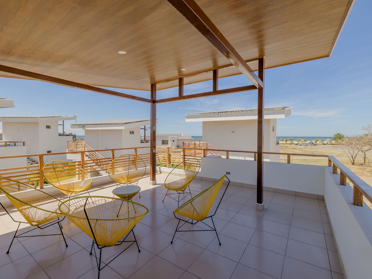 A covered balcony with yellow wire chairs and a table overlooks white buildings and a distant body of water under a clear sky.