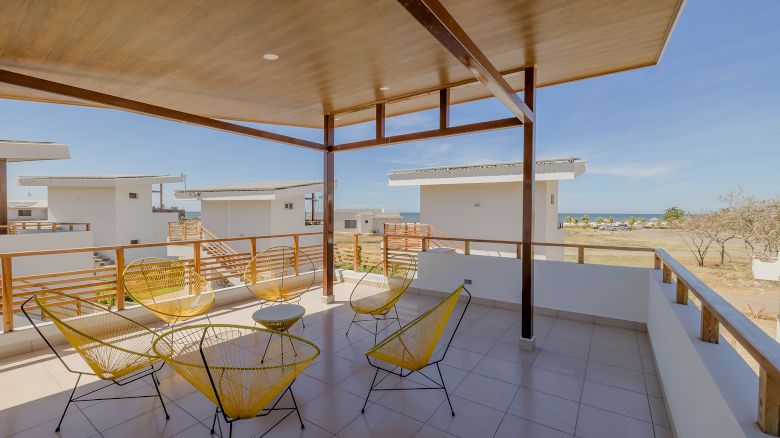 A covered balcony with yellow wire chairs and a table overlooks white buildings and a distant body of water under a clear sky.