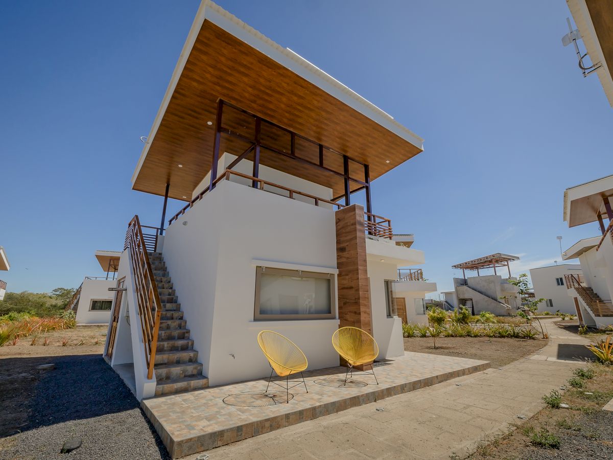 The image shows a modern two-story house with an outdoor staircase, two yellow chairs on the patio, and clear skies in the background.