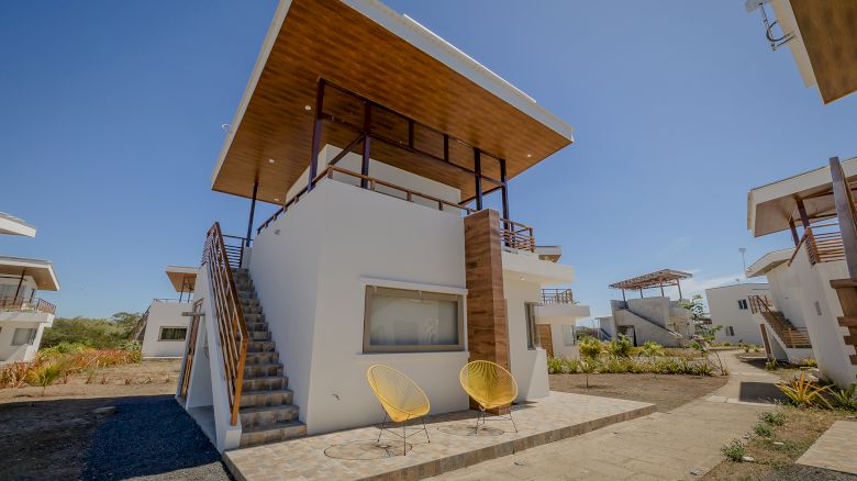 The image shows a modern two-story house with an outdoor staircase, two yellow chairs on the patio, and clear skies in the background.