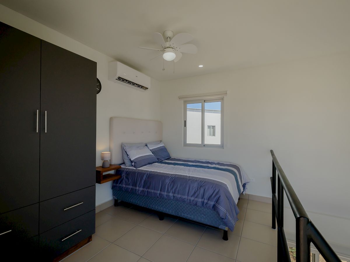 A bedroom with a bed, blue bedding, a nightstand, a wardrobe, air conditioner, a window, a ceiling fan, and a railing on the right side.