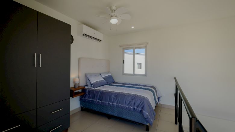 A bedroom with a bed, blue bedding, a nightstand, a wardrobe, air conditioner, a window, a ceiling fan, and a railing on the right side.