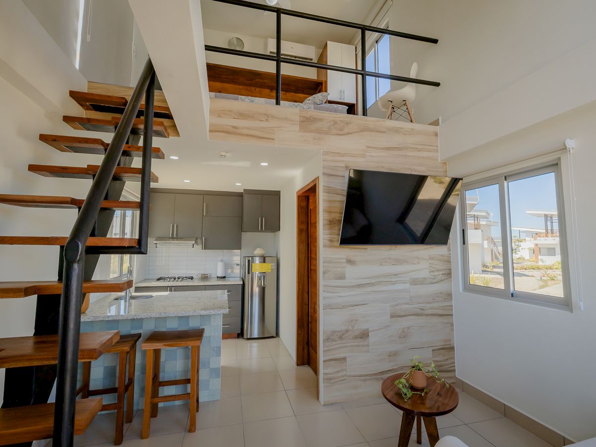 A modern loft apartment with a wooden staircase, kitchen, TV, and a small table with fruit. Bright natural light pours through the window.