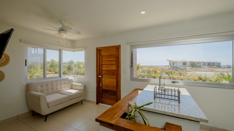 A cozy room features a sofa, wooden door, and large windows with a scenic view. A countertop island with a plant adds to the inviting atmosphere.