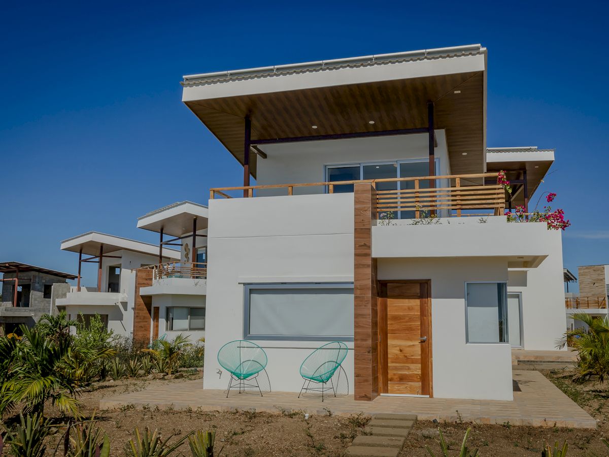 A modern two-story house with large windows, wooden accents, a balcony, and outdoor seating, surrounded by plants and clear blue skies.