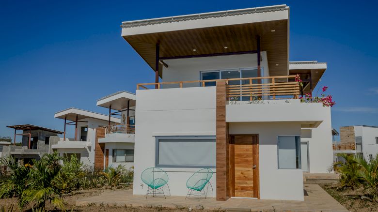 A modern two-story house with large windows, wooden accents, a balcony, and outdoor seating, surrounded by plants and clear blue skies.