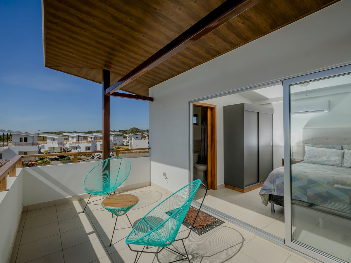 A modern balcony with two turquoise chairs and a small table, adjacent to a bedroom with a bed, and large glass sliding doors.