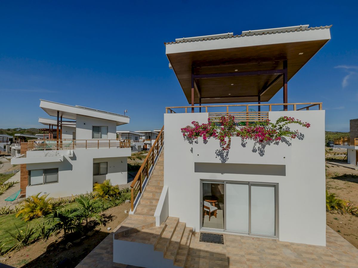 The image shows a modern two-story house with a rooftop patio, stairs, and flowering plants on the balcony, set in a residential area on a clear day.