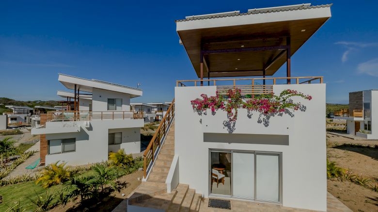 The image shows a modern two-story house with a rooftop patio, stairs, and flowering plants on the balcony, set in a residential area on a clear day.