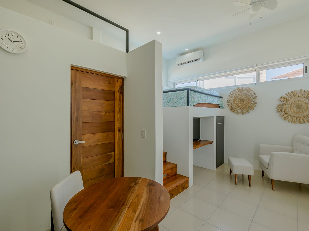 A modern, compact room with a wooden door, a round wooden table, white chairs, a cozy bed, and decorative wall pieces, plus natural lighting.