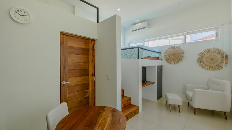 A modern, compact room with a wooden door, a round wooden table, white chairs, a cozy bed, and decorative wall pieces, plus natural lighting.