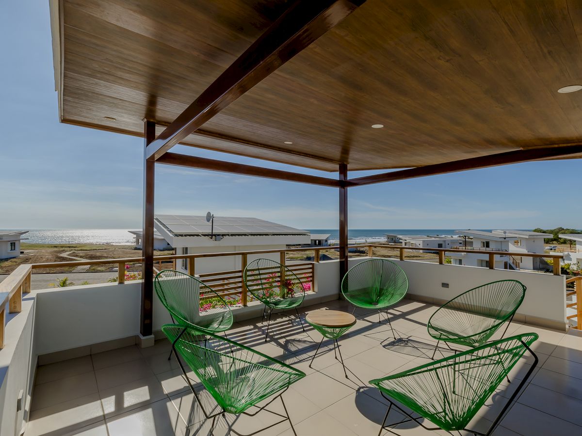 An outdoor patio with green wire chairs and a table, overlooking rooftops and the ocean, covered by a wooden ceiling.