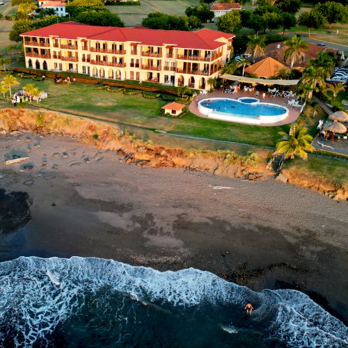 An aerial view of a large building with a red roof, a circular pool, and surrounding greenery, located on the edge of a sandy beach with waves.