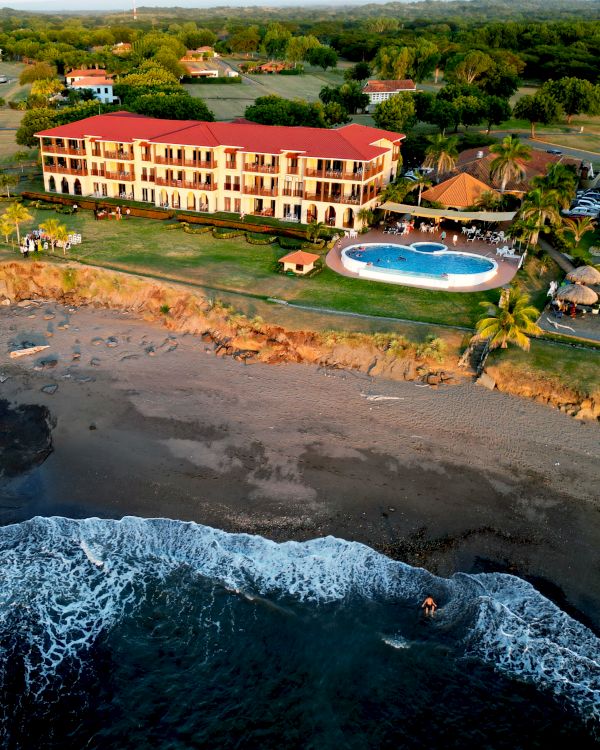 An aerial view of a large building with a red roof, a circular pool, and surrounding greenery, located on the edge of a sandy beach with waves.