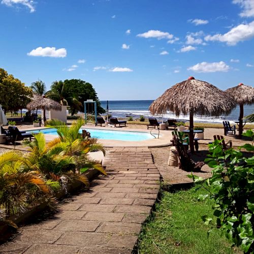 A tropical resort with a pathway leading to a pool surrounded by palm trees and sun umbrellas, overlooking a calm ocean under a partly cloudy sky.