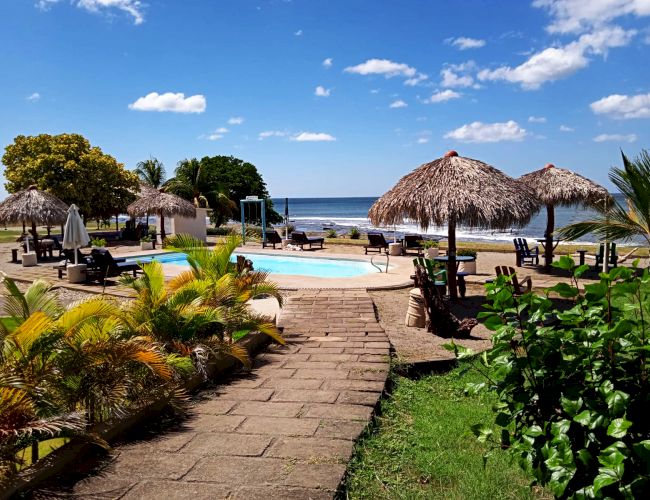 A tropical resort with a pathway leading to a pool surrounded by palm trees and sun umbrellas, overlooking a calm ocean under a partly cloudy sky.