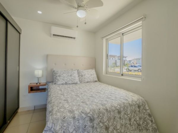 A small bedroom with a bed, nightstand, lamp, window, air conditioner, ceiling fan, and sliding door closet. The bed has a patterned comforter.